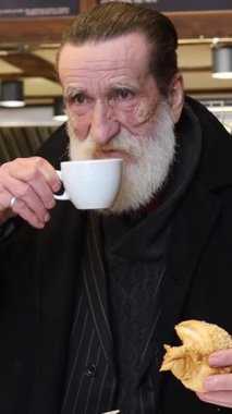 Sakallı yaşlı adam sıcak bir kafe ortamında çay ve pastanın tadını çıkarıyor. Dikey video.