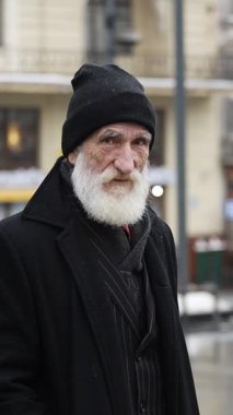 Beyaz sakallı, siyah giyinmiş yaşlı bir adam canlı bir şehir ortamında duruyor.