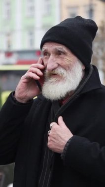 Siyah bereli ve kalın paltolu yaşlı bir adam dışarıda telefonuyla sohbet ediyor..