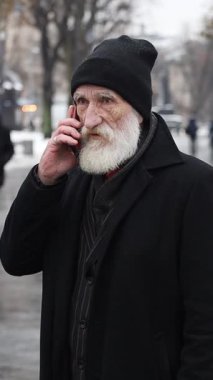 Siyah paltolu ve bereli yaşlı adam şehir ortamında yürürken akıllı telefon kullanıyor. Dikey video.