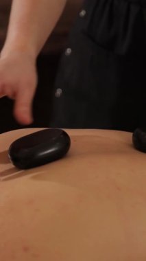 Lüks kaplıcalarda sıcak taş masajı yaptıran bir kadın. Rahatlatıcı ve sakin bir atmosferde rahatlatıcı bir terapi. Dikey video.