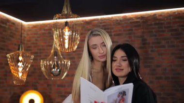 Gelin ve asistan, gelinlikleri katalogdan seçerken bir yandan da düğün salonunda tuğla duvar arkasında dikiliyorlar. Şık bir düğün salonu.