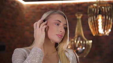 Lüks beyaz elbiseli çekici, orta yaşlı bir sarışının yakın plan portresi. Sarışın, aynada Swarovski taşlarıyla küpe deniyor..