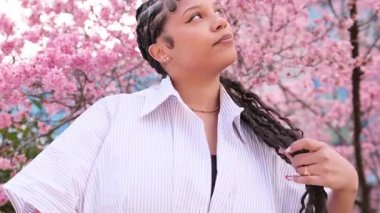 Siyah tenli beyaz gömlekli Afro-Amerikalı bir kadının portresi kameraya poz veren pembe sakura 'nın arka planına karşı. Avrupalı esmer kadın pembe sakura çiçeğinin arka planında.