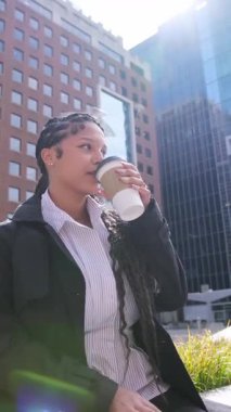 Genç Afrikalı Amerikalı kadın güneşli bir günde gökdelenlerin arka planında kahve içiyor. Kadın sıcaklığın tadını çıkarıyor, dışarıda kahve molası veriyor.