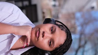 Dikey video. Açık alanda bulanık bir arka planda, kulaklık takmış koyu tenli bir kadının yakın plan portresi. Güzel bir Afrikalı Amerikalı kadının yüzü.