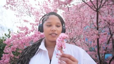 Kulaklıklı, pembe sakura arka planında telefonu olan pozitif bir Amerikalı kadının çok güzel bir videosu. Güzellik kavramı, doğanın keyfi ve sağlıklı yaşam tarzı..