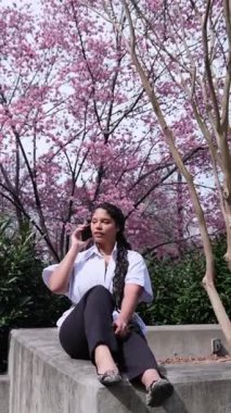 Beyaz tişörtlü, pembe sakura arka planda oturan, telefonda konuşan Afro-Amerikalı kadın. Baharda şehirde esmer tenli Amerikalı kadın