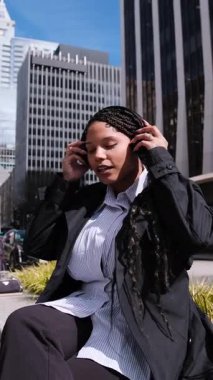 Güzel bir Afro-Amerikalı kadın, büyük bir şehirde sokakta otururken müzik dinliyor. Güneşli bir günde kadın dışarıda kulaklıkla müzik dinliyor.