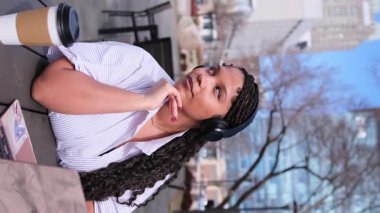 Dikey video. Bir kafede kahve masasında oturan, kulaklıklı, gülümseyen esmer bir kadının portresi. Baharda açık havada güzel bir Afrikalı Amerikalı kadın.