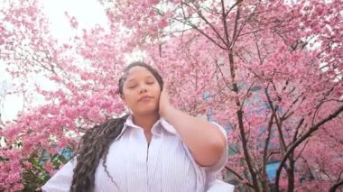 Beyaz gömlekli Afro-Amerikalı bir kadının portresi pembe sakura 'nın arka planına karşı, poz vererek, güzelliğin tadını çıkararak. Avrupalı kadın pembe sakura çiçeğinin arka planına karşı.