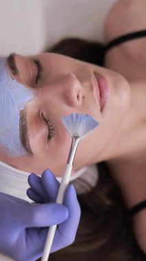 Bir güzellik uzmanı genç bir kadına yüz maskesi takar. Güzellik salonunda gençleştirme prosedürleri. Dikey video.