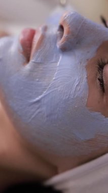 Genç bir bayana yüz maskesi takan bir güzellik uzmanı. Güzellik salonunda gençleştirme prosedürleri. Profesyonel yüz bakımı. Dikey video
