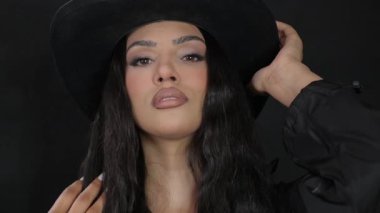 Siyah şapkalı ve kıyafetli şık bir kadın koyu bir arka plana karşı özgüven ve zarafet yayıyor..