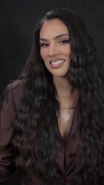 Uzun dalgalı saçlı, kendine güvenen bir iş kadını koyu bir arka plana karşı resmi giyinirken sıcak bir şekilde gülümser..