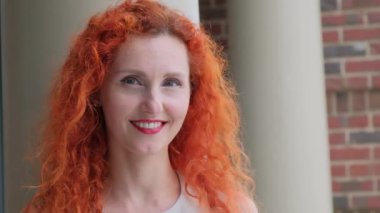 Güzel kıvırcık kızıl saçlı neşeli genç bir kadın zarif bir açık havada parlak bir şekilde gülümsüyor.