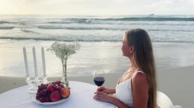 Kadın sahilde güzel bir gün batımında şarap ve taze meyvenin tadını çıkarıyor..