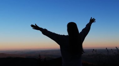 Bir kadının silueti, kollarını mükemmel bir çerçeveye uzatarak dağlarda nefes kesici bir günbatımının arka planına karşı ayakta duruyor.