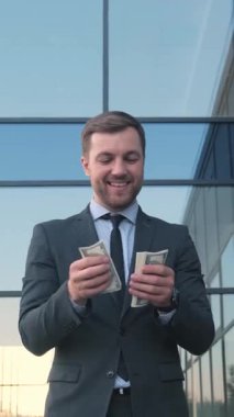 Keskin takım elbiseli neşeli ve başarılı bir işadamı yansıtıcı bir ofis binasının önünde yığınla para sayıyor.