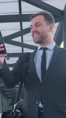 Takım elbiseli profesyonel bir adam havaalanı terminalinde akıllı telefonunu kullanırken geniş bir gülümseme sergiliyor.