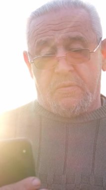 Açık hava ortamında, gözlüklü yaşlı bir adam dikkatle akıllı telefon cihazına bakar.