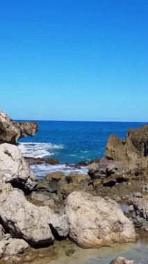 Güneşli bir gökyüzünün altında dalgaların kıyı şeridine çarpmasıyla kayalık bir plajın güzelliğini hissedin. Okyanusların güçlü hareketlerini gösterin. Dikey video