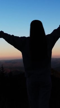 Gün batımında tepede duran, özgürlüğünü ve doğayla bağlantısını kucaklayan bir kadının zarif silueti. Dikey video.