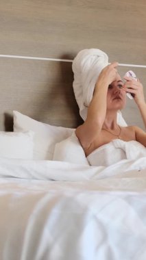 Bir kadının yatağa sarılıp, yumuşak ışık ve sessiz yansımayla yıkandığı huzurlu bir an. Dikey video.