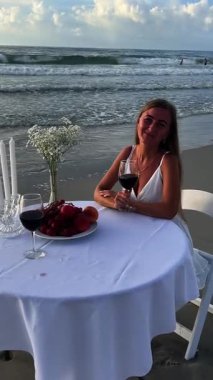 Sahilde kırmızı şarapla yemek yiyen bir kadın, çeşitli meyveler ve gün batımında okyanusun büyüleyici manzarası..