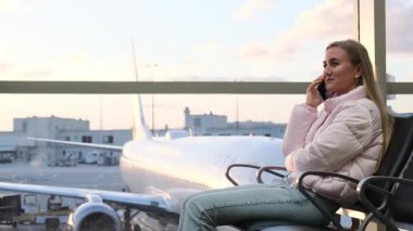 Pembe ceketli genç bir kadın havaalanı salonunda otururken bir telefon görüşmesi yapıyor.