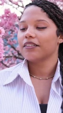 Afrikalı bir Amerikalı kadın, renkli bir bahar günü, canlı pembe kiraz çiçekleriyle çevrili doğanın güzelliğini neşeyle kucaklıyor. Dikey video
