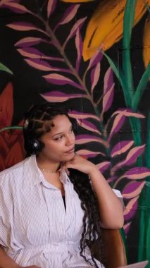 Zarif bir Afrikalı genç Amerikalı kadın şehir ortamında renkli parlak bir duvar resminin yanında güvenle poz verirken müzikten zevk alıyor. Dikey video