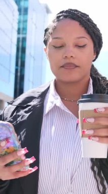 İş kıyafetleri içinde Afro-Amerikalı bir kadın canlı bir şehir ortamında açık havada kahve içerken akıllı telefonunu kullanıyor. Dikey video