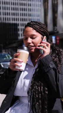 Kendinden emin genç bir Afrikalı Amerikalı profesyonel canlı bir şehir ortamında telefonla konuşurken kahveden zevk alır. Dikey video