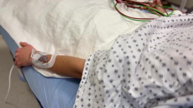 Burada bir hastane ortamında serumlu ve çeşitli izleme cihazlı bir hastanın kolunun yakından görüntüsü var.