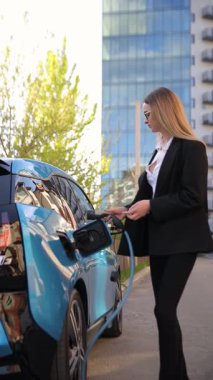 Güneşli otoparkta genç bir kadın elektrikli arabayı şarj ediyor. Dikey video.