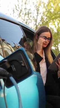 Gözlüklü bir iş kadını elektrikli arabanın içindeki tabletten şarj olurken mesaj okuyor. Dikey video.