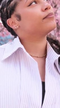 Beyaz gömlekli Afro-Amerikalı kadın, baharda çiçek açan pembe sakuranın arka planına karşı güven içinde poz veriyor. Dikey video