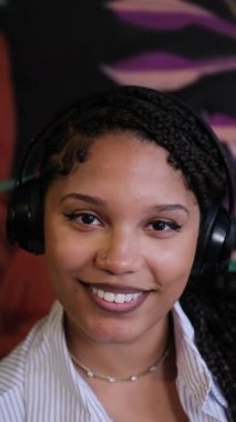 Güzel kar beyazı gülüşlü, kulaklık takan ve pozitif ışıldayan Afro-Amerikan bir kadının yakın çekimi. Dikey video