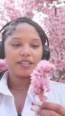 Kulaklık takan ve telefon tutan olumlu bir Amerikalı kadın çiçek açan pembe sakuradan, güzelliği, doğayı ve sağlıklı yaşam tarzını kucaklamaktan zevk alır. Dikey video