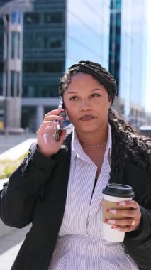 Kendine güvenen Afrikalı Amerikalı iş kadını, arka planda yüksek binalar olan bir şehir caddesinde kahve içerken telefonla konuşuyor. Dikey video