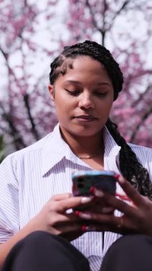 Koyu tenli Amerikalı bir kadın bahar boyunca şehir dışında sakura ağaçlarının altında akıllı telefonunu kullanarak oturur. Dikey video