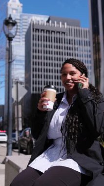 Takım elbiseli gülümseyen Afro-Amerikalı bir kadın açık havada, modern şehir ortamında kahveyi seviyor. Dikey video