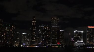 Gece Miami şehir merkezinin etkileyici bir manzarası. Işıklı binalar ve şehrin yansımaları suyun üzerinde parıldıyor..