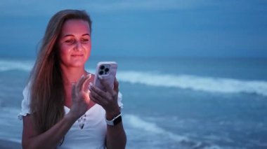 Alacakaranlıkta sahilde duran bir kadın, telefonuna gülümsüyor. Arka planda okyanus dalgalarıyla.. 