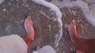 Romantik çift gün batımında kumsalda yalınayak yürüyor. Nazik dalgalar ve altın ışıkla yakın çekimde..