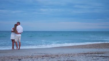 Genç çift kıyı şeridinde sarılıyor, dalgalar ve yumuşak akşam ışığı ile çevrili.. 