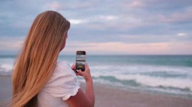 Uzun sarı saçlı genç bir kadın gün batımında okyanusun fotoğrafını çekiyor.. 