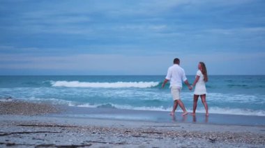Birbirine aşık bir çift, dalgalar ve yumuşak akşam ışıklarıyla çevrili kıyı şeridi boyunca yürür. Güzel bir tatil..