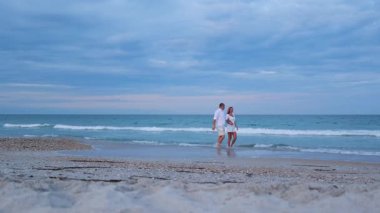 Denizin kenarında yürüyen çift, sakin atmosferde sohbet ediyor ve gülümsüyor.. 